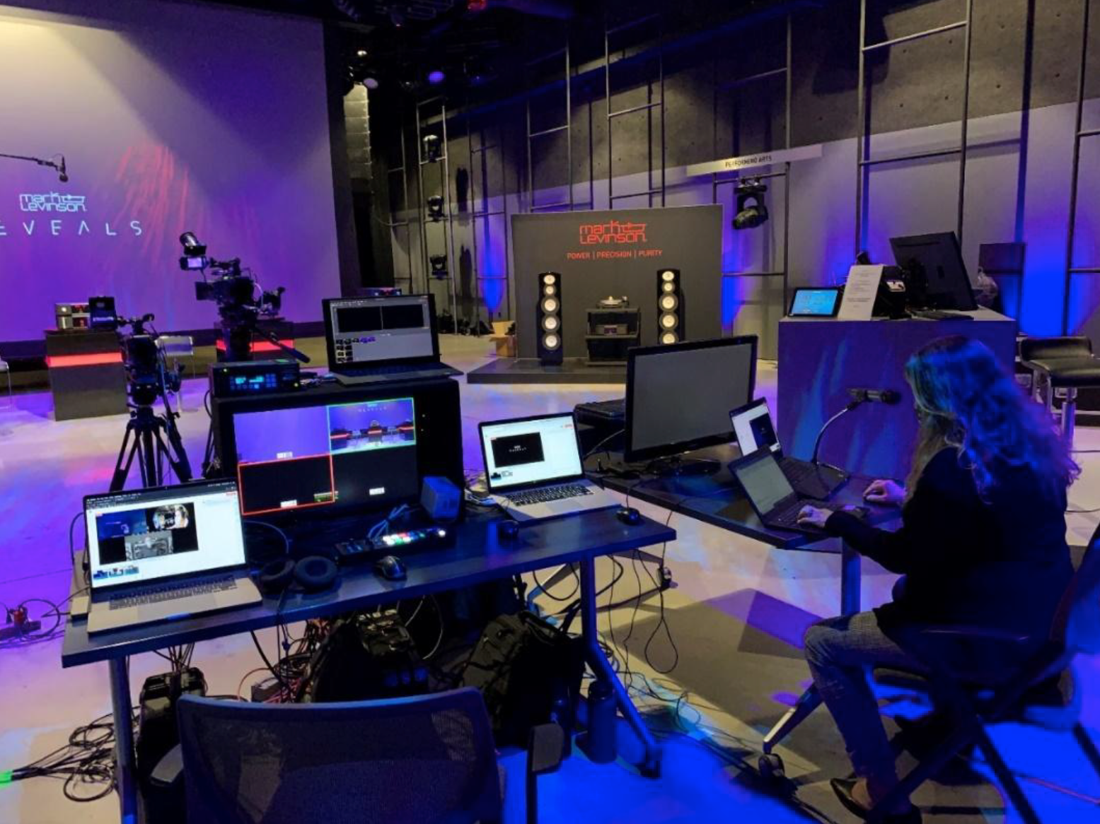 A person sits at a desk with several monitors, laptops, and video equipment in a studio with cameras, lights, and a stage setup reading Webster Reveals in the background.