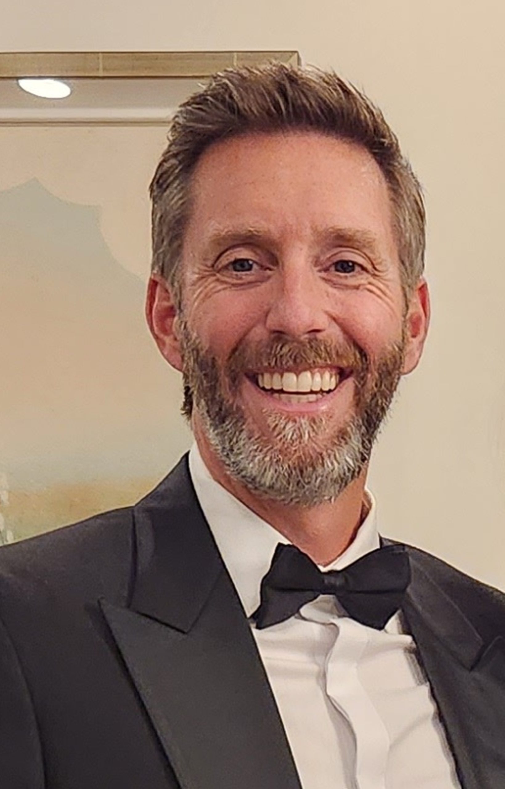 A man with short brown hair and a beard smiling broadly is dressed in a black tuxedo white shirt and black bow tie The background is softly lit with neutral colors
