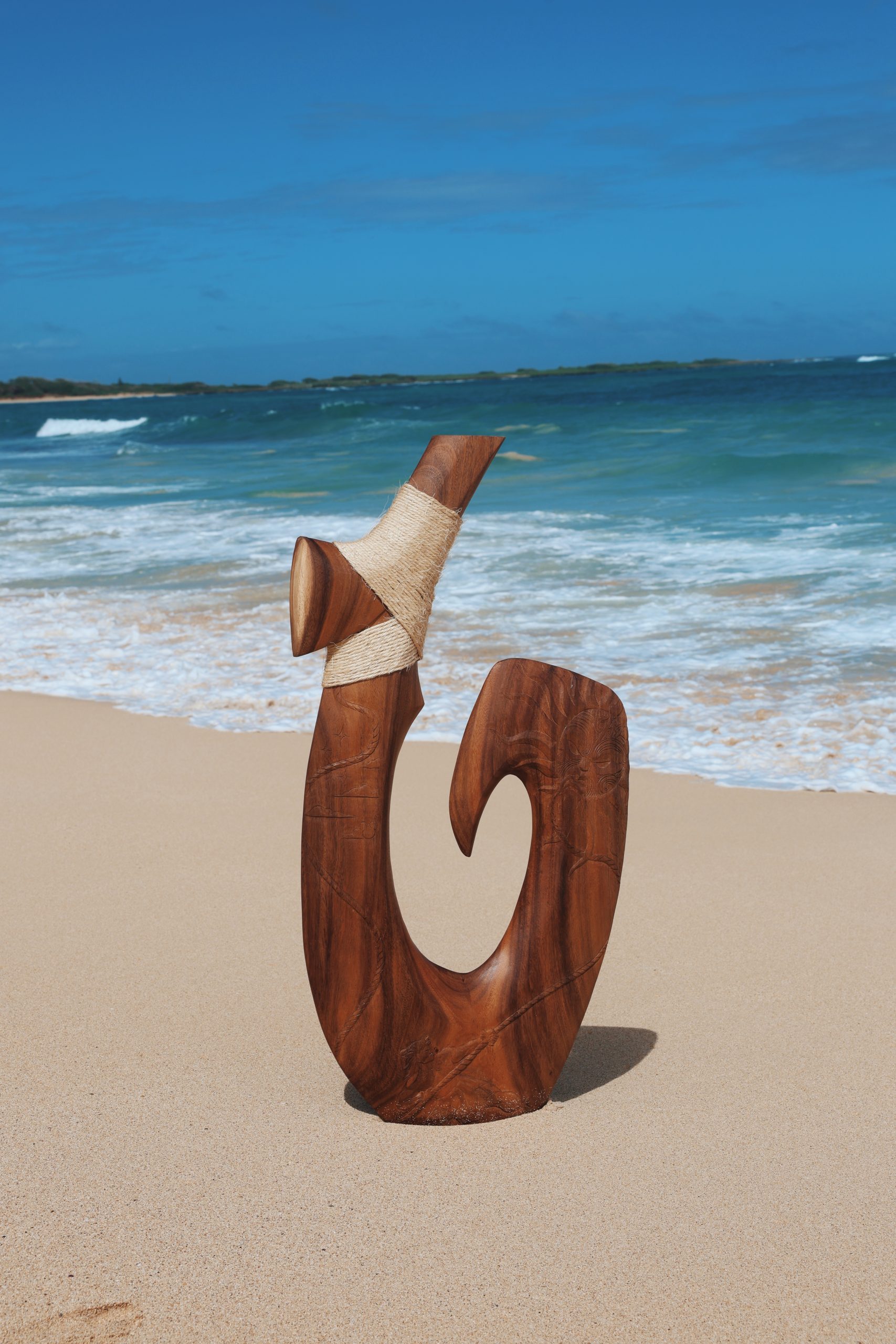 A carved wooden sculpture with a spiral design wrapped partly in rope stands upright on a sandy beach with ocean waves and a blue sky in the background