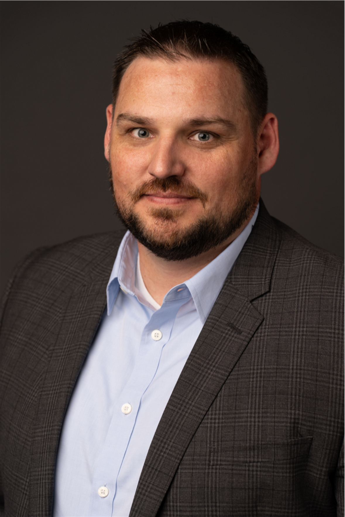 A man with short dark hair a trimmed beard and light skin wears a gray plaid suit jacket over a light blue dress shirt posing against a dark backgroundready to share his bright ideas