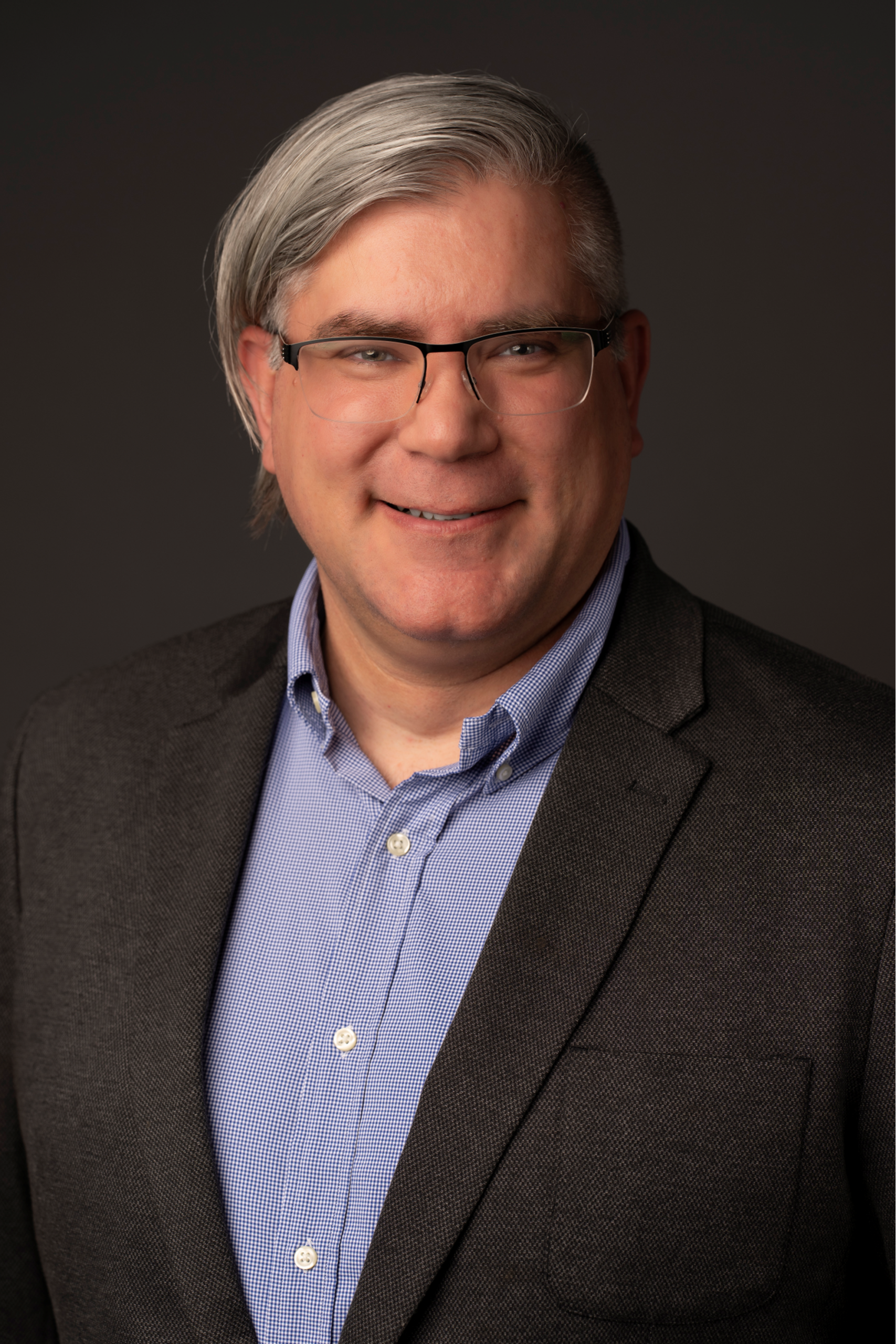 A man with gray hair wearing glasses a blue checkered shirt and a dark blazer smiles at the camera against a dark backgroundradiating confidence and bright ideas