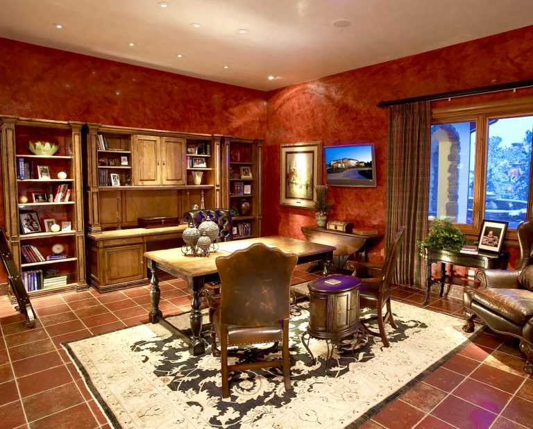 A warm elegant home office with red textured walls wooden bookshelves a desk with ornate chairs a patterned rug and a window letting in natural light Decor includes plants framed photos and vases