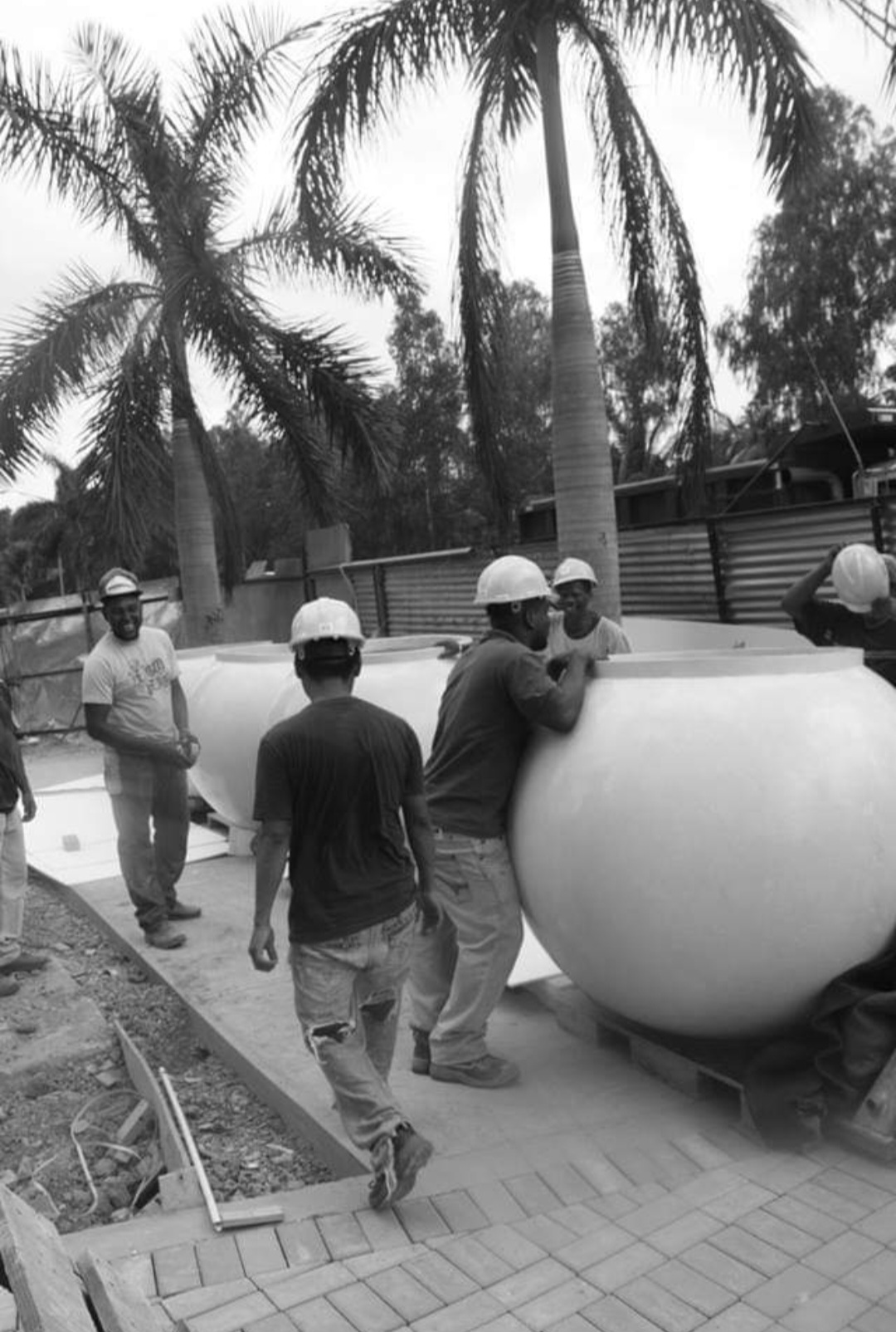 A group of construction workers wearing hard hats move a large round planter near palm trees at a construction site Built across borders some workers are lifting while others stand by