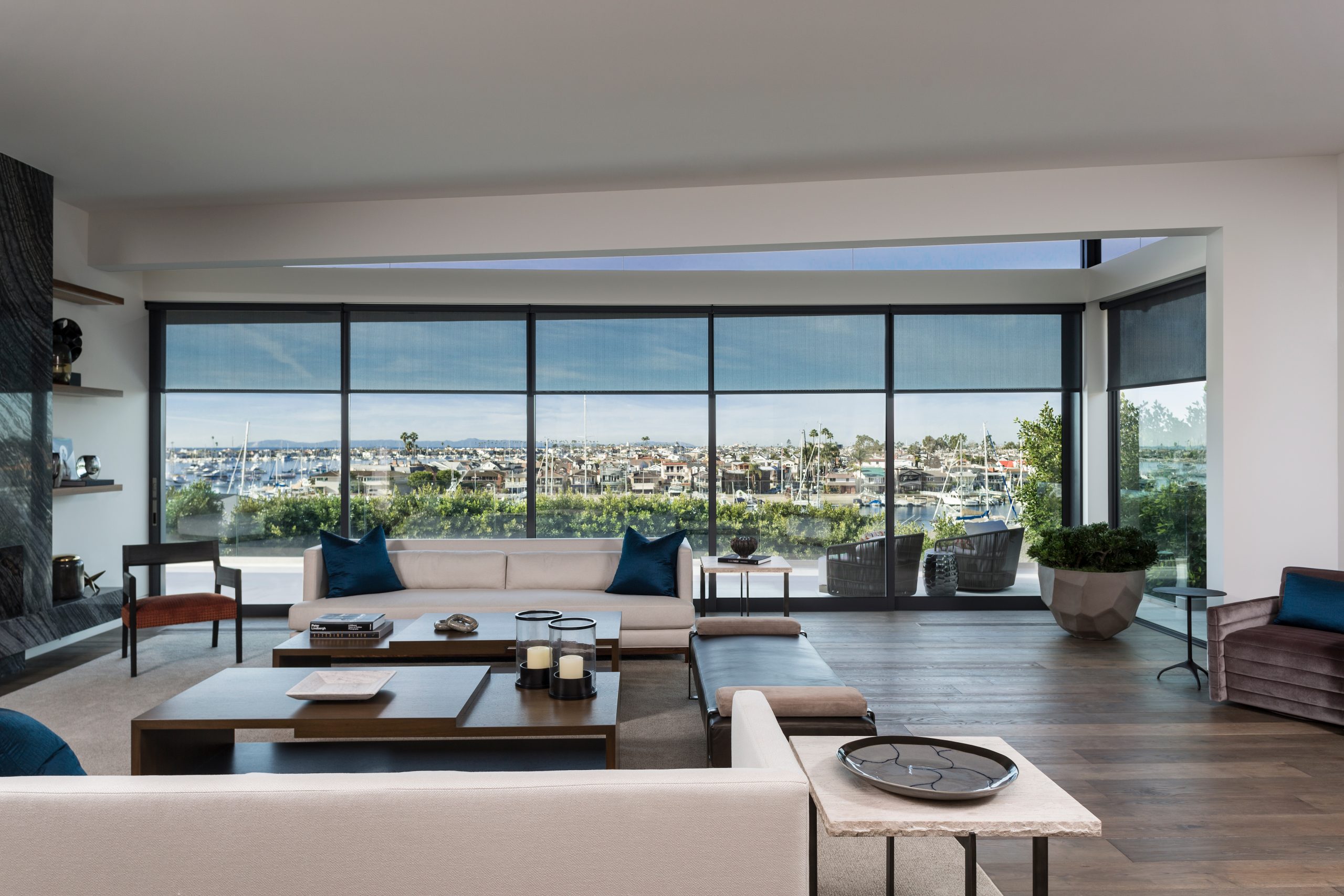 Modern living room with large windows offering a view of a marina and boats furnished with neutral toned sofas dark wood tables decorative items and savant smart home features plus a potted plant by the window