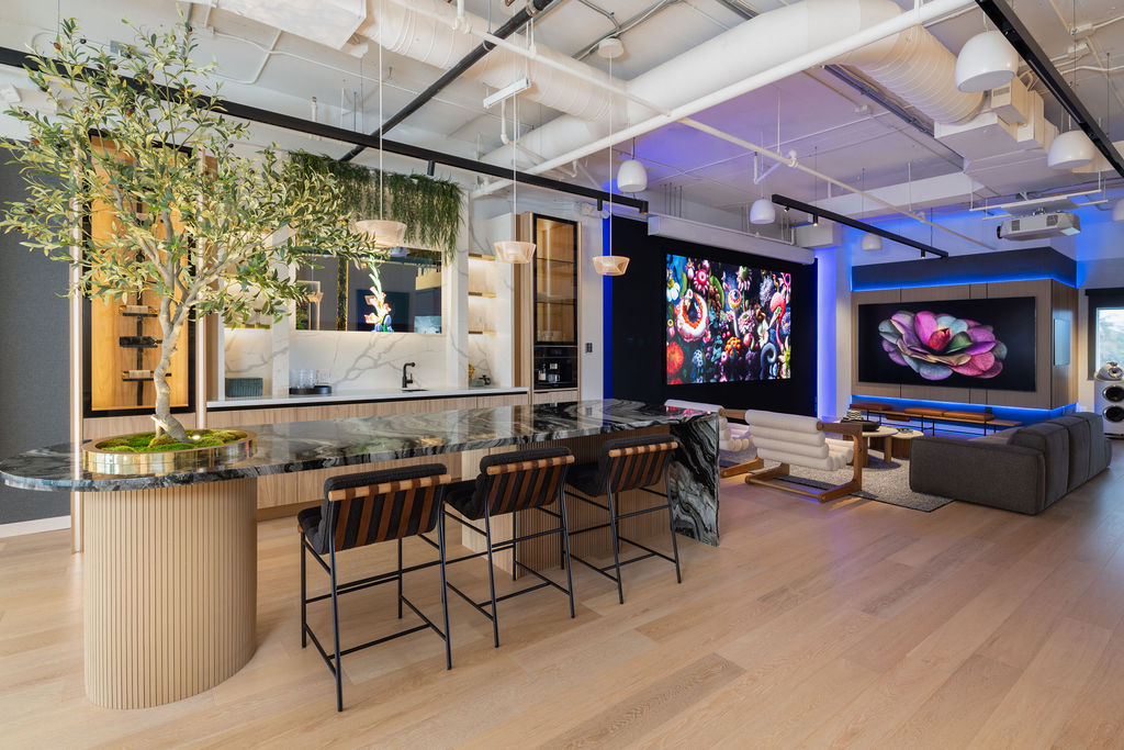 Modern open concept living space at the showkase experience center featuring a marble kitchen island bar stools lush indoor plants and a cozy lounge area facing two digital screens Exposed ceilings and warm wood flooring by lk & associates