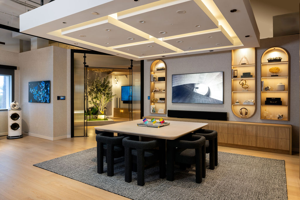 Modern living room by lk & associates with a large wooden table and eight black chairs on a gray rug built in shelves wall mounted tv and soft lighting Glass doors open to a room with a tree and ambient lightdiscover more at our experience center