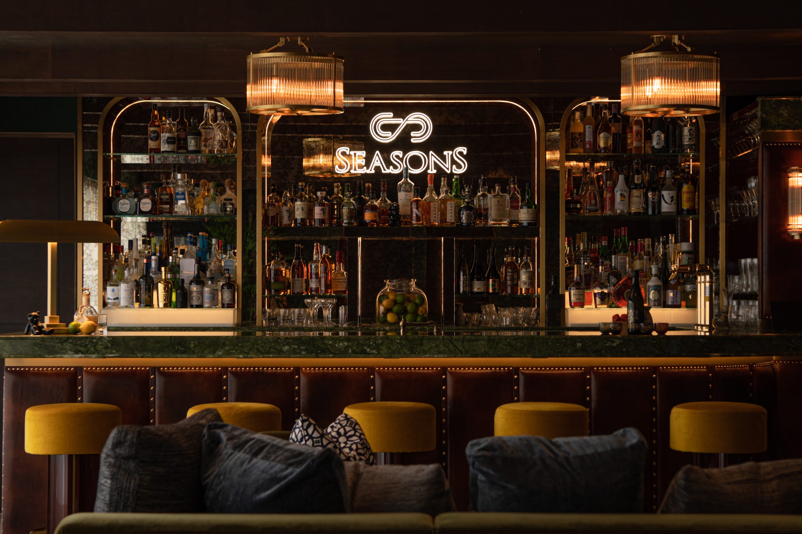 Elegant bar interior with cushioned stools bottles lined on mirrored shelves two large pendant lights and a glowing seasons sign above the bar Inspired by japan the warm lighting creates a cozy and inviting atmosphere