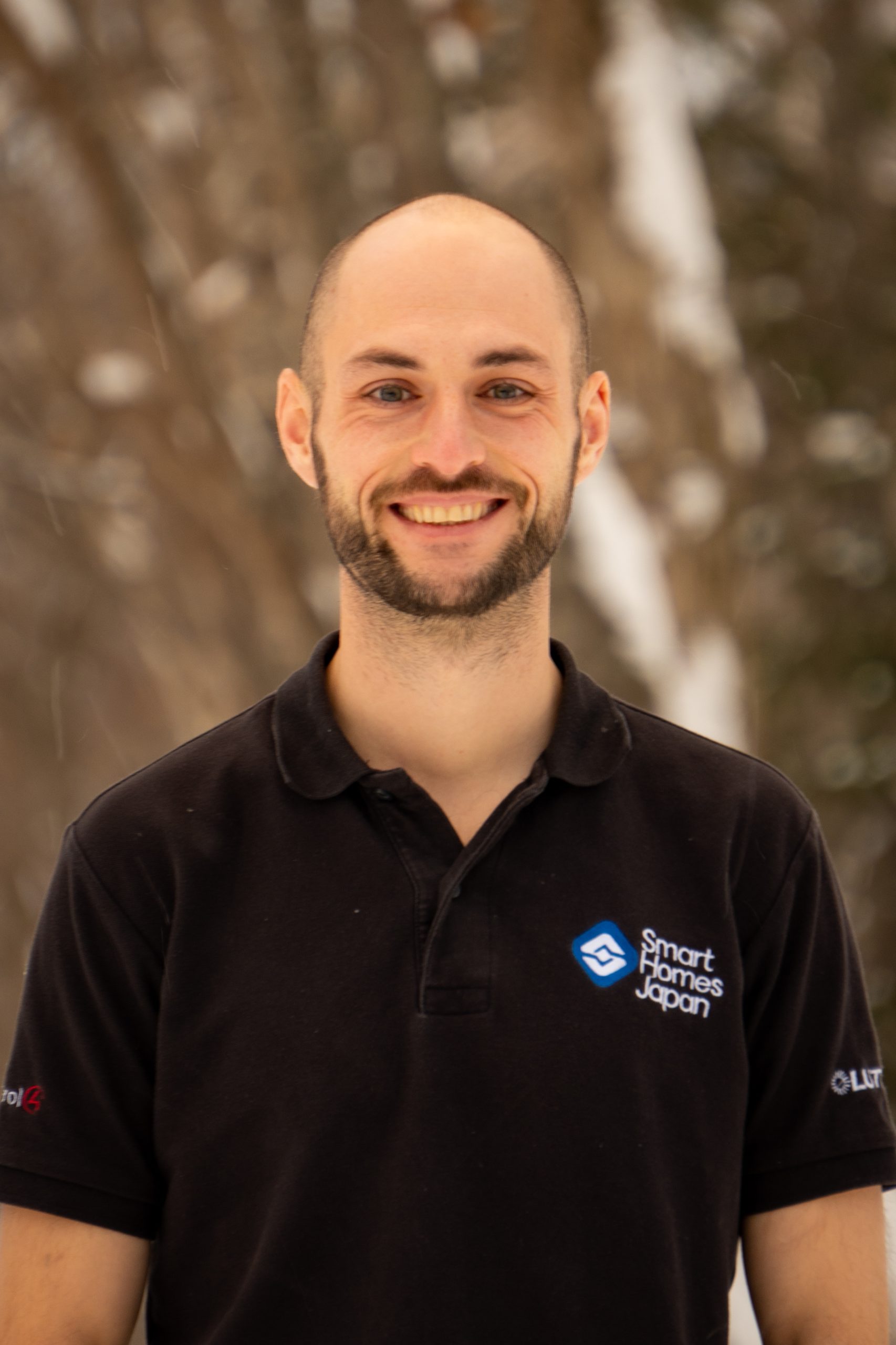 A smiling man with a bald head and beard stands outdoors in winter He wears a black polo shirt with smart homes japan and logos representing smart home integration Snowy trees are blurred in the background