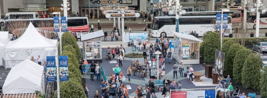 A busy outdoor event area with many people walking among tents, booths, and displays—some featuring Performance Home products—surrounded by greenery and parked buses in the background. Signs and banners are visible throughout the scene.