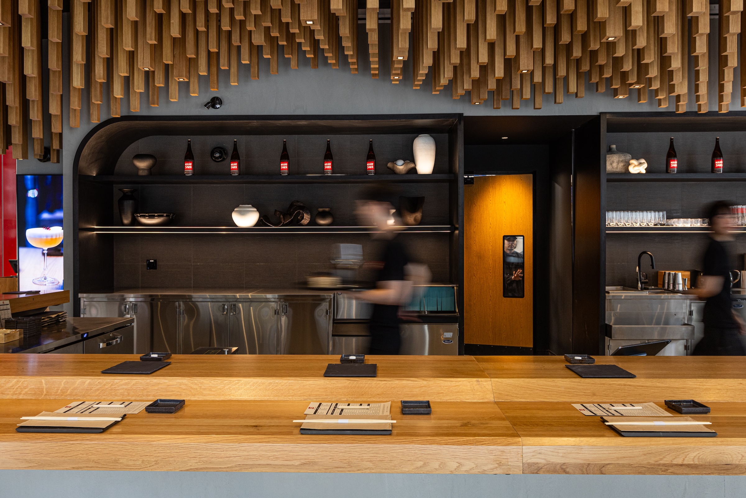 A modern sushi bar with a wooden counter stylish hanging lighting fixtures and ceiling decor shelves with bottles and bowls and two blurred staff members moving behind the counter A modern sushi bar with a wooden counter stylish hanging lighting fixtures and ceiling decor shelves with bottles and bowls and two blurred staff members moving behind the counter Lighting big ideas with small fixtures from usai lord abbett hello nori & 9 w 57th