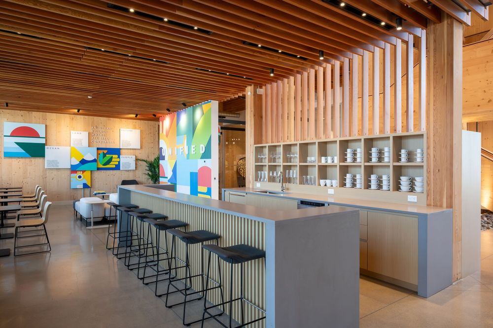 Modern café with a long gray bar lined with black stools, open wooden shelving holding white dishes, light wood walls, and colorful geometric artwork on the walls. Ceiling features wooden slats and recessed lighting.