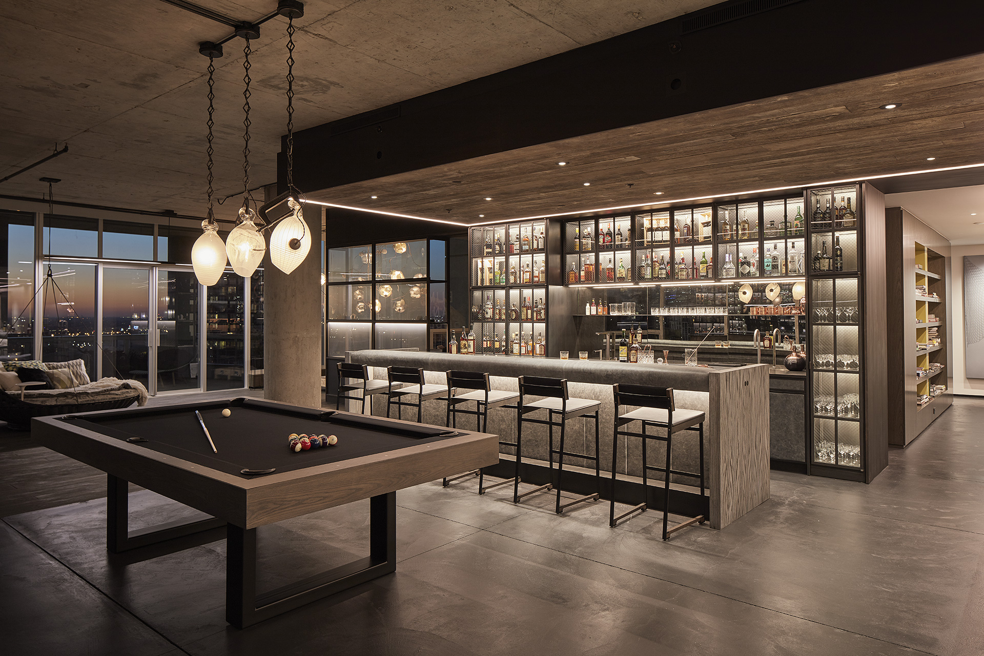 Modern home bar and lounge with a pool table sleek lit bar counter with high stools illuminated shelves displaying bottles large windows and warm ambient lighting City skyline visible through windows in the evening Modern home bar and lounge with a pool table sleek lit bar counter with high stools illuminated shelves displaying bottles large windows and warm ambient lighting City skyline visible through windows in the evening Luxury integrated technologies its lit