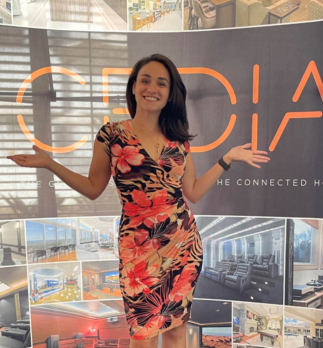 A woman in a floral dress stands smiling with arms outstretched in front of a display board featuring cedia and images of modern home interiors showcasing the innovation behind luxury integrated technologies lit A woman in a floral dress stands smiling with arms outstretched in front of a display board featuring cedia and images of modern home interiors showcasing the innovation behind luxury integrated technologies lit Luxury integrated technologies its lit