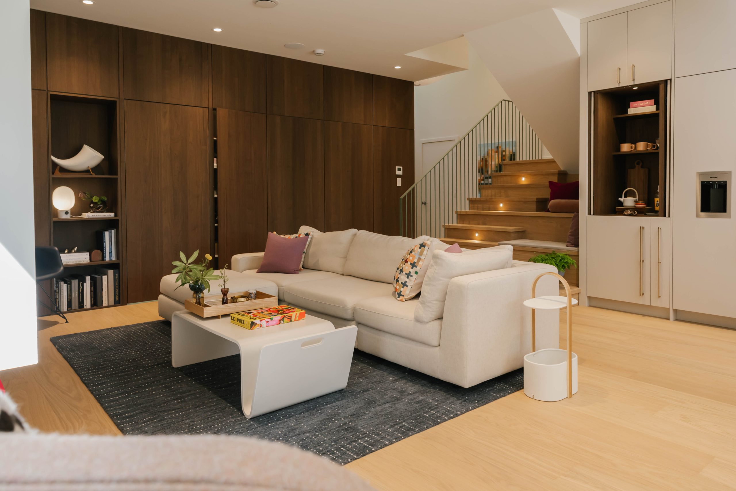 Modern living room with a beige sofa colorful pillows coffee table bookshelf and staircase with lights Wooden wall and floor white cabinets plants and decor create a cozy stylish atmosphere Modern living room with a beige sofa colorful pillows coffee table bookshelf and staircase with lights Wooden wall and floor white cabinets plants and decor create a cozy stylish atmosphere 2026 technology designer magazine performance home award winners