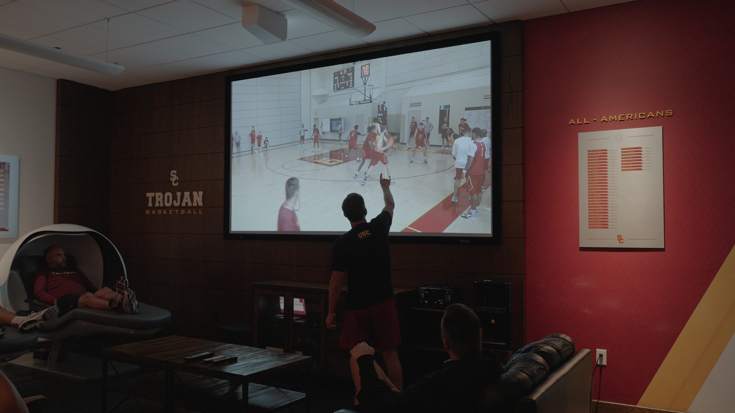 A coach points at a large screen showing a basketball practice while players and staff watch in a usc basketball meeting room 2026 technology designer magazine performance home award winners